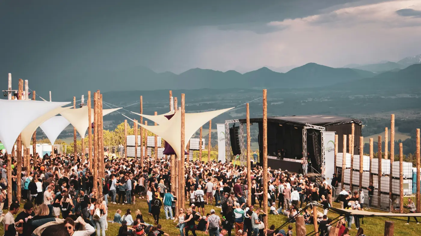 Dağlara bakan bir yamaçta, beyaz gölgeliklerin altında sahnenin önünde dolaşan festival kalabalığı