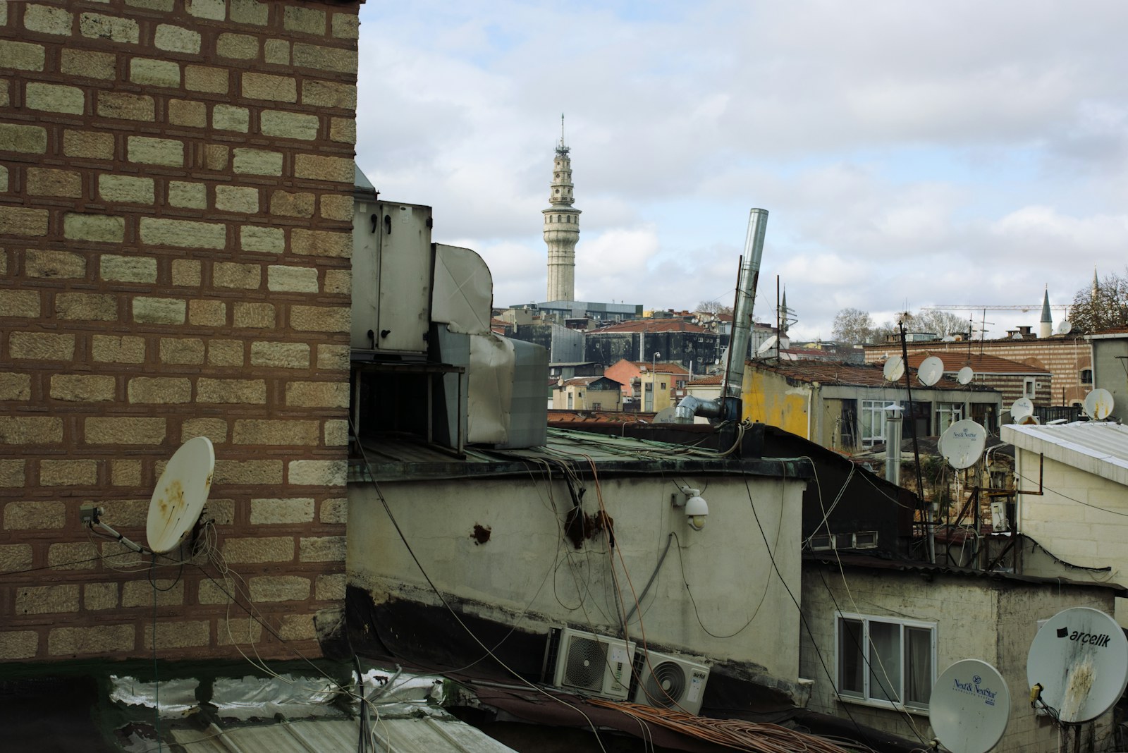 Çatılar ve uydu antenleri üzerinden görünen şehir; ötede bir kule ve bulutlu gökyüzü