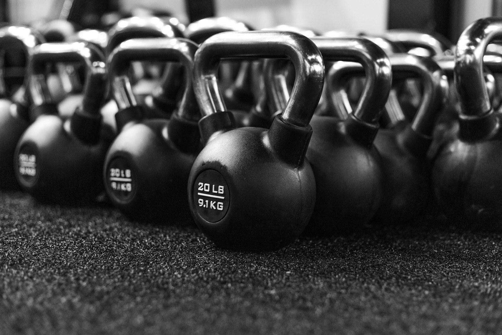 Kauçuk zeminde yan yana duran dökme demir kettlebell’lerin siyah beyaz fotoğrafı; ağırlıkları libre ve kilogramla yazılı