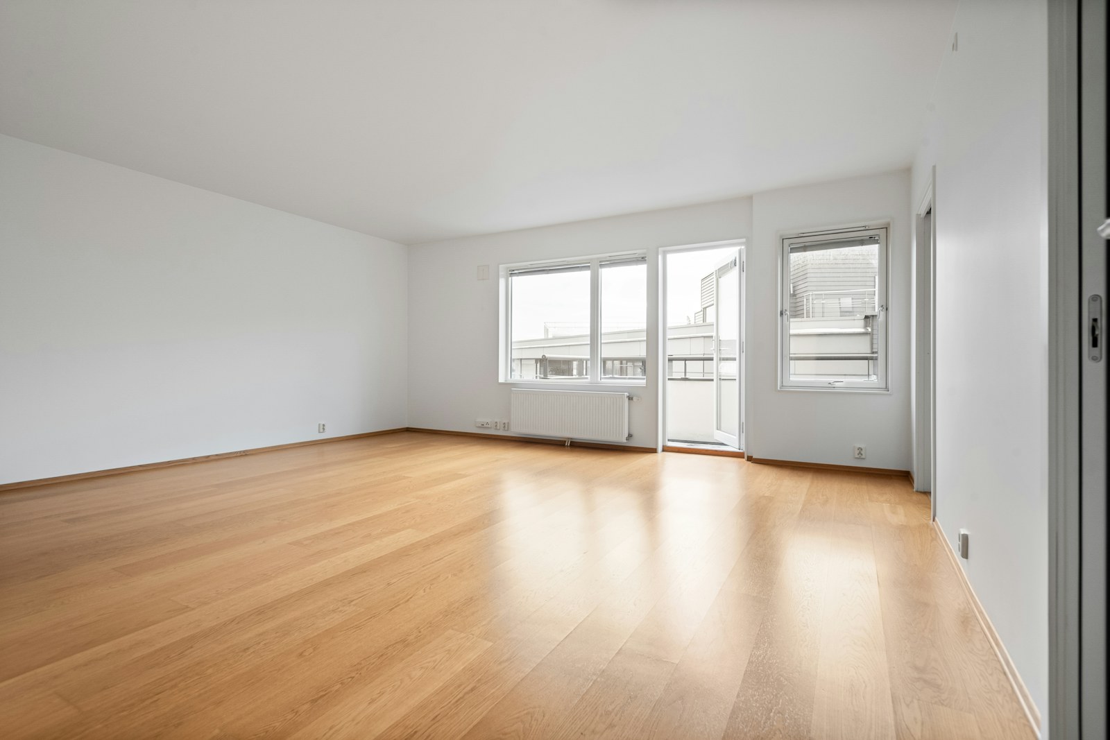 Parke zeminli, beyaz duvarlı boş bir daire odası; pencerelerden ve balkon kapısından gün ışığı giriyor.